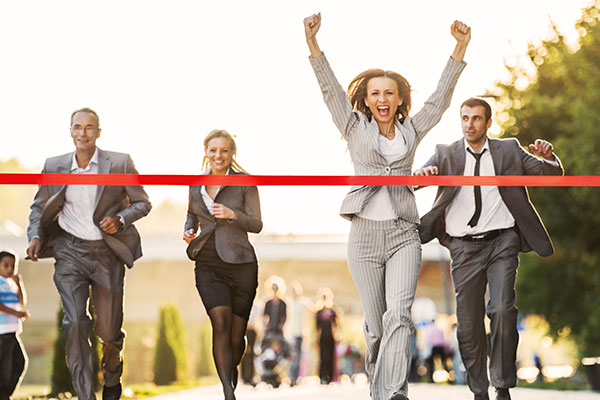 Two men and two women in business attire running toward a finish line made of red tape