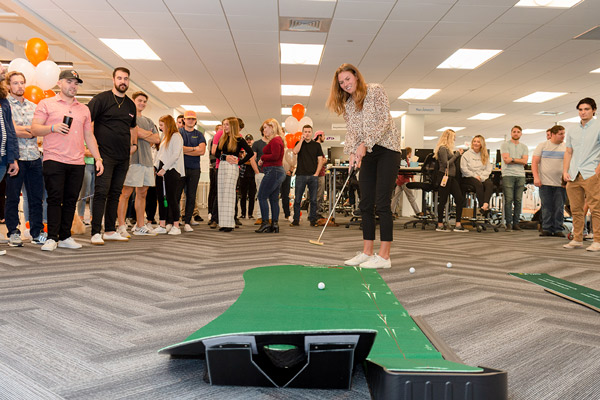 Female employee playing mini golf at the office while coworkers look on